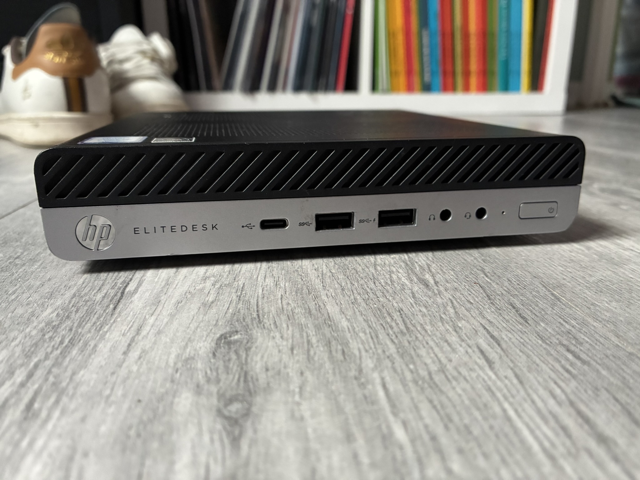 Front panel of an HP EliteDesk mini PC on a wooden floor, showing the USB-C port, two USB-A ports, headphone jacks and the power button.